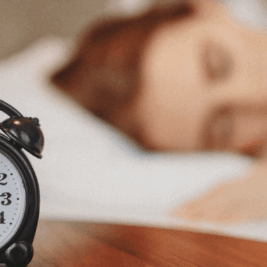 blurry image of a girl sleeping peacefully in the background, with a focused alarm clock in the foreground, symbolizing the connection between rest and mental health