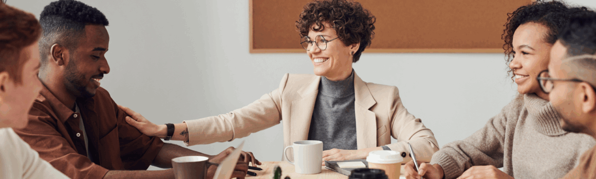 employees at work, sitting around a table having a meeting while smiling, promoting a healthy work environment focused on mental health